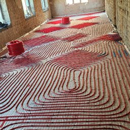 Envi-Technik Przemysław Ruszczyński - Czerwone rury ogrzewania podłogowego ułożone na folii w surowym wnętrzu. Widoczne rolki rur. W tle okna i surowa ściana z cegły.