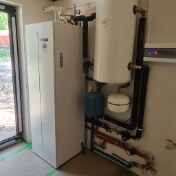 Envi-Technik Przemysław Ruszczyński - Biała pompa ciepła York z zbiornikiem wody i orurowaniem w kotłowni z pianką izolacyjną na suficie. Widoczne rury instalacji grzewczej.