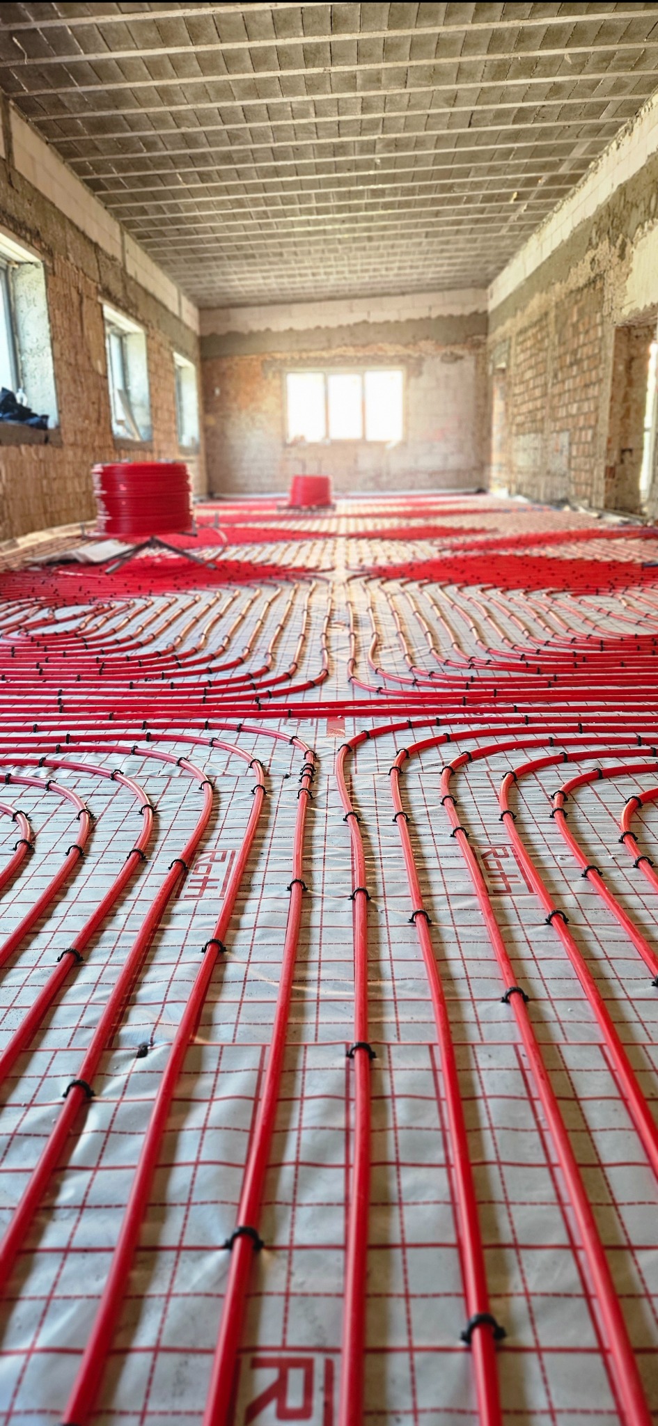 Czerwone rury ogrzewania podłogowego ułożone na białej macie w surowym wnętrzu z oknami. Widoczne zwoje rur i surowy betonowy strop.