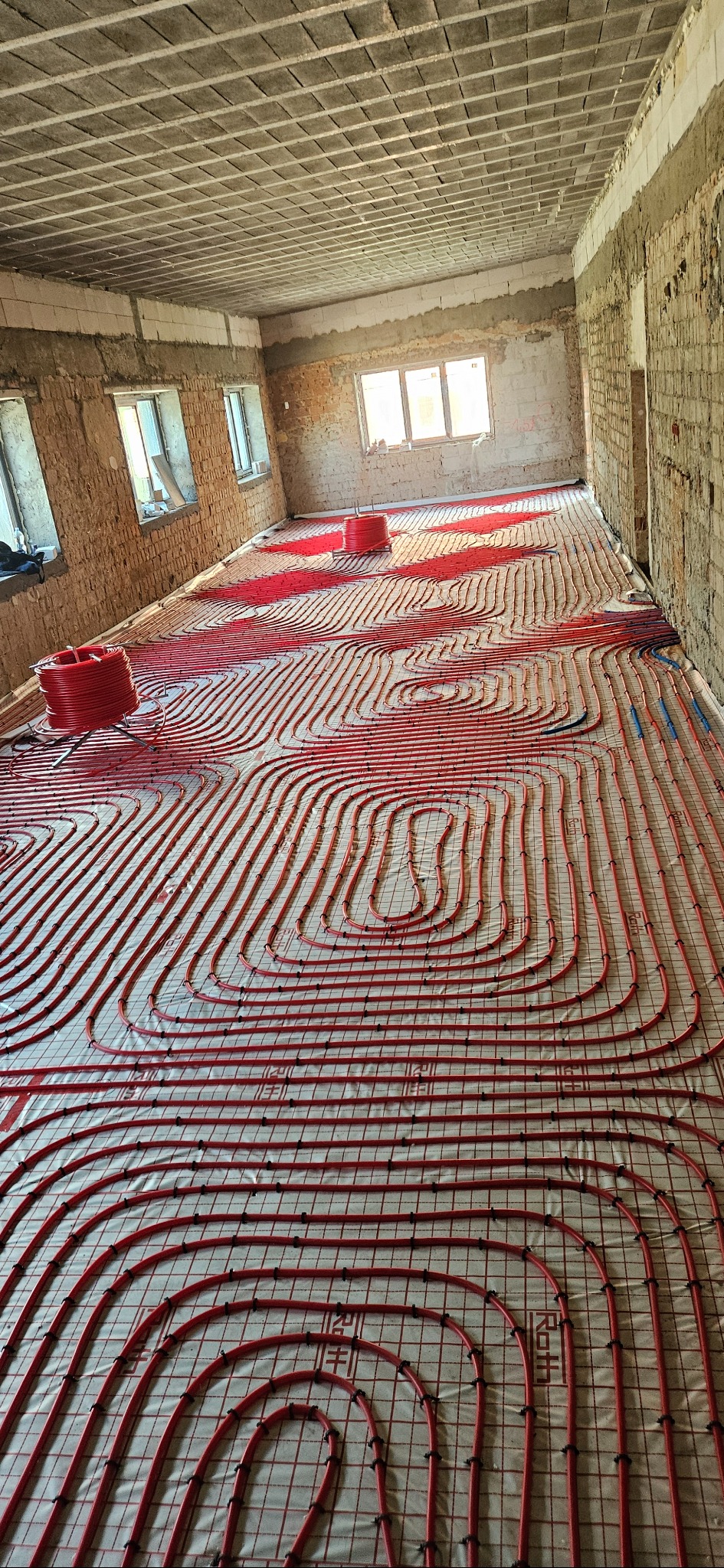 Czerwone rury ogrzewania podłogowego ułożone na folii w surowym wnętrzu. Widoczne rolki rur. W tle okna i surowa ściana z cegły.