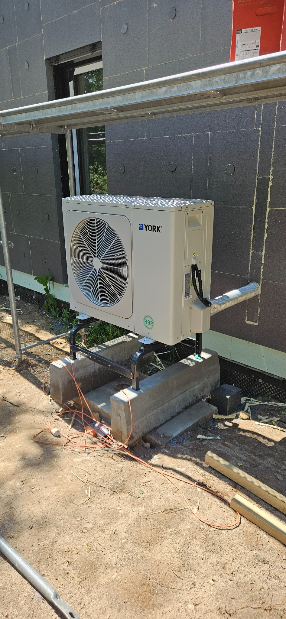 Biała pompa ciepła marki York zamontowana na betonowym fundamencie przy budynku z szarą elewacją. Widoczne rusztowanie i kabel zasilający na ziemi.