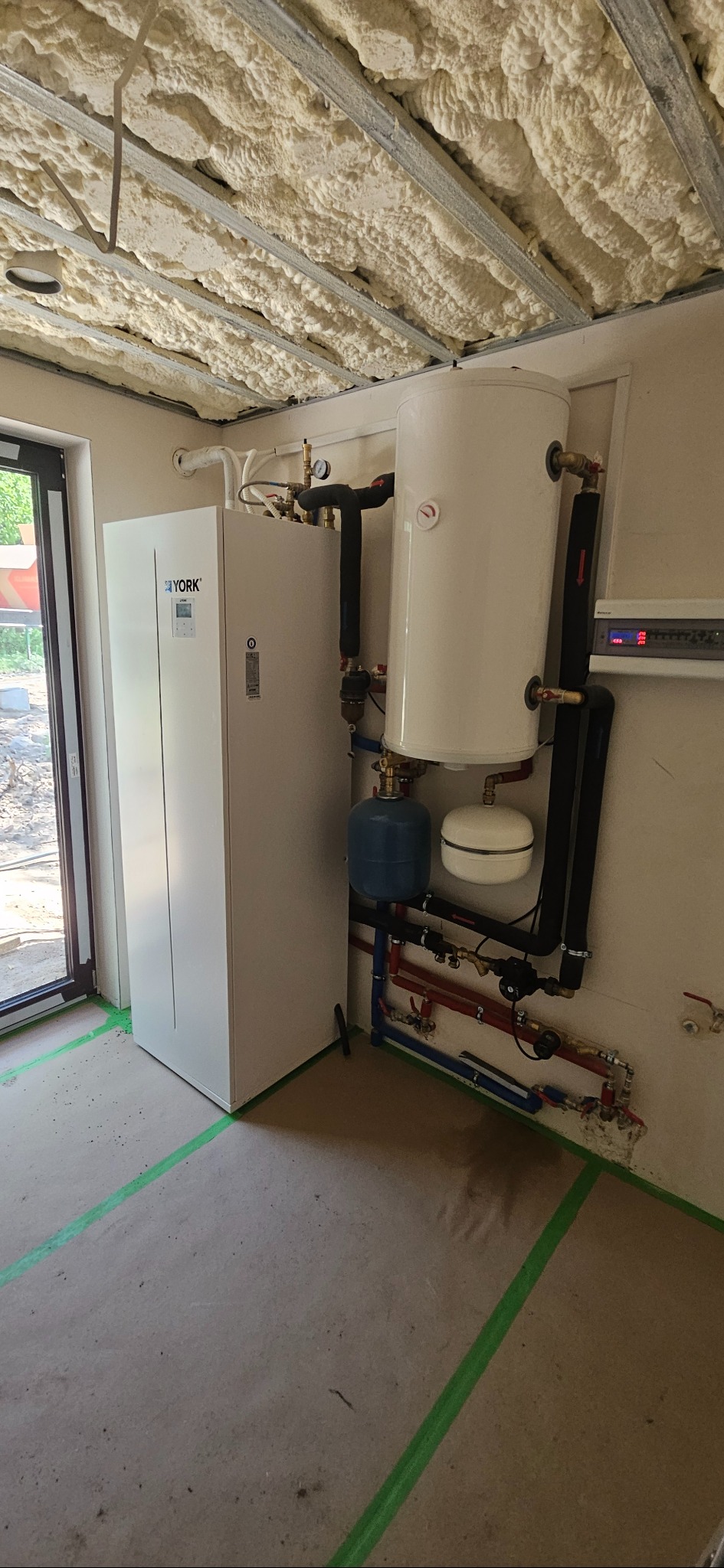 Biała pompa ciepła York z zbiornikiem wody i orurowaniem w kotłowni z pianką izolacyjną na suficie. Widoczne rury instalacji grzewczej.