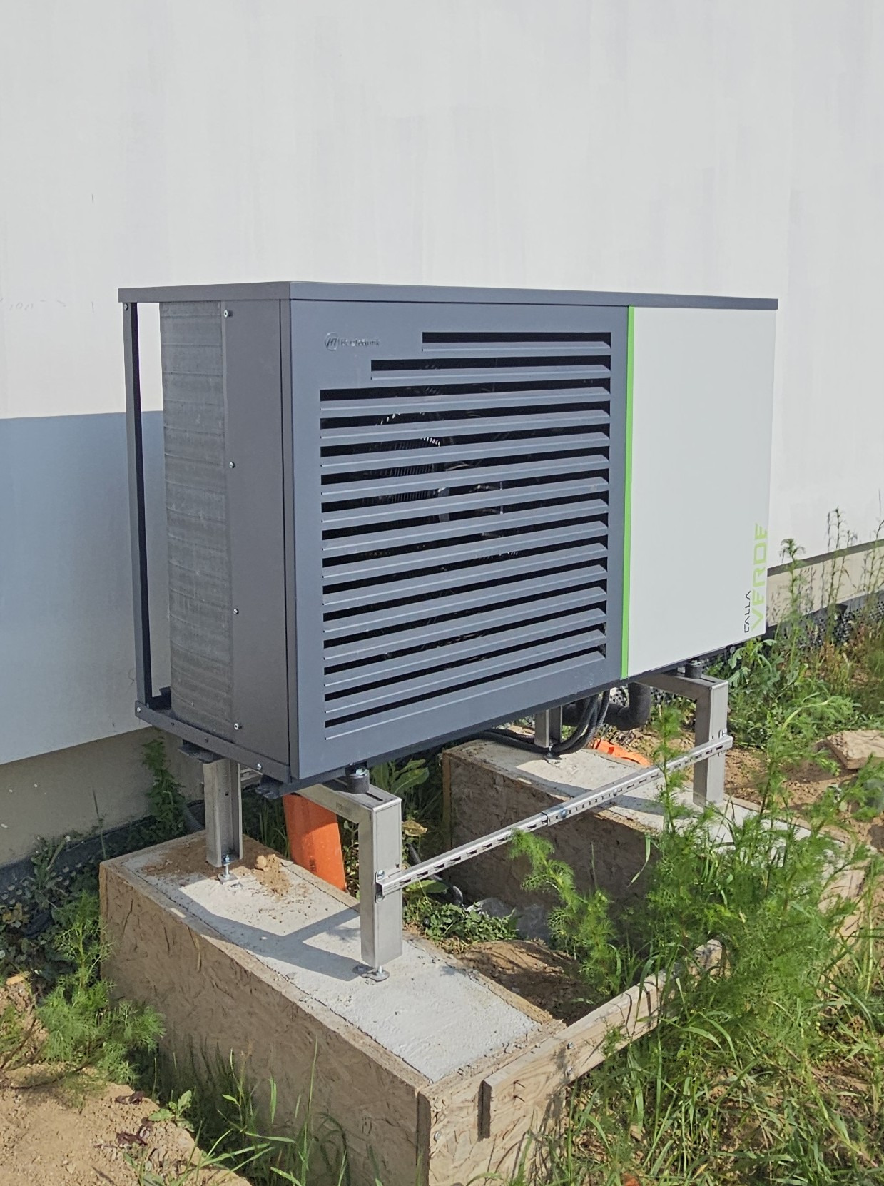 Szara pompa ciepła zamontowana na metalowej konstrukcji na betonowym fundamencie przy białej ścianie budynku, otoczona zielenią. Widoczne detale montażu.