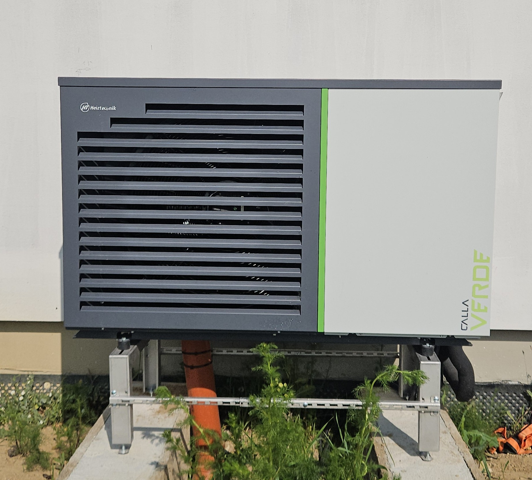 Szara pompa ciepła Heitztechnik Calla Verde na betonowej podstawie, z widocznymi rurami i zielenią w tle, zamontowana przy jasnej ścianie budynku.
