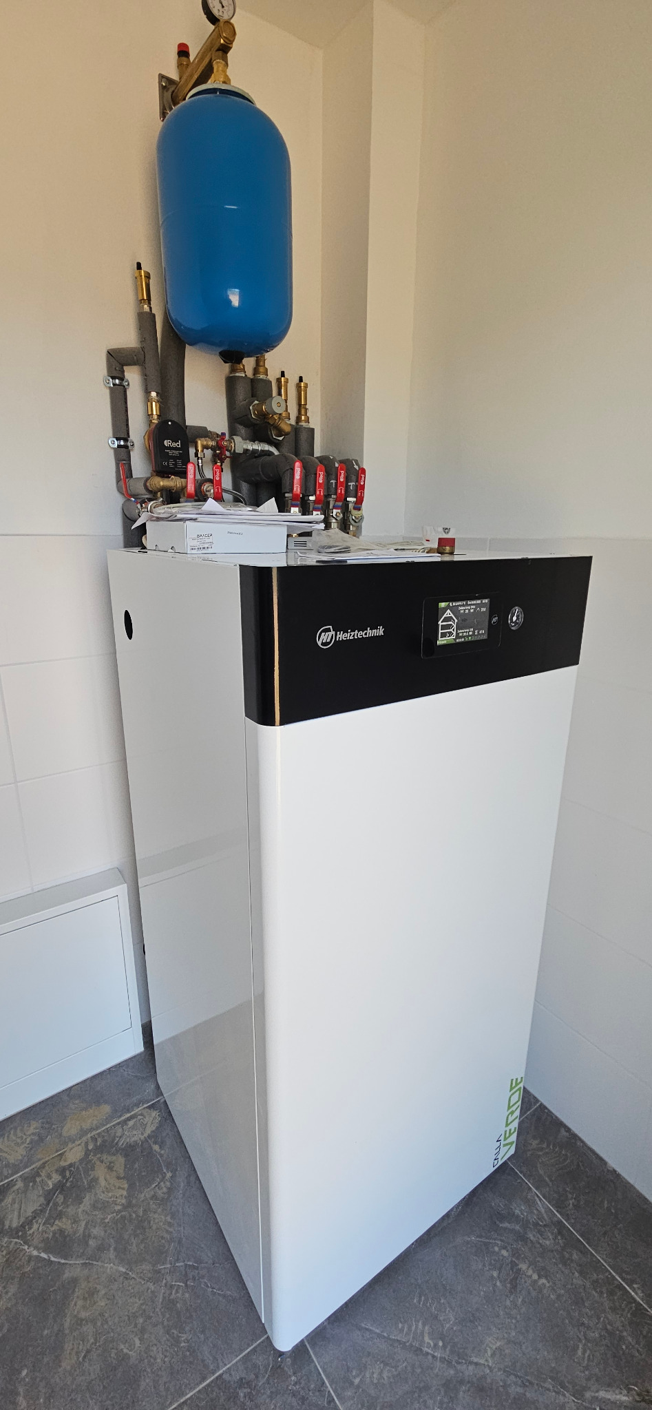 Nowoczesny, biały piec gazowy Heiztechnik z czarnym panelem sterowania i niebieskim zbiornikiem wyrównawczym, zamontowany w rogu pomieszczenia. Widoczne rury i zawory.