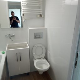 Marek Szpakowski - Nowoczesna, biała łazienka po remoncie: podwieszana toaleta, szafka pod umywalką i grzejnik drabinkowy. W lustrze odbija się osoba robiąca zdjęcie.