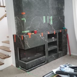Marek Szpakowski - Obudowa kominka z ciemnego marmuru w trakcie montażu, zabezpieczona taśmą malarską. Widoczne narzędzia i poziomica. Schody w tle.