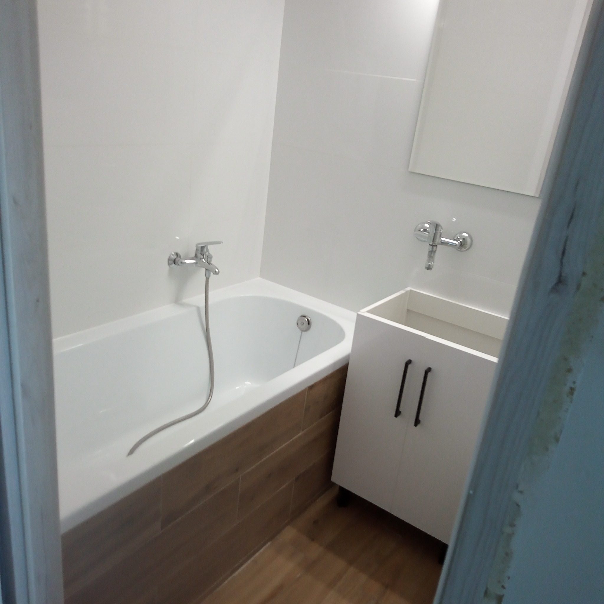 Biała wanna z obudową imitującą drewno, umywalka z szafką i lustro w wykończonej łazience. Bateria wannowa i umywalkowa chromowana. Podłoga drewnopodobna.