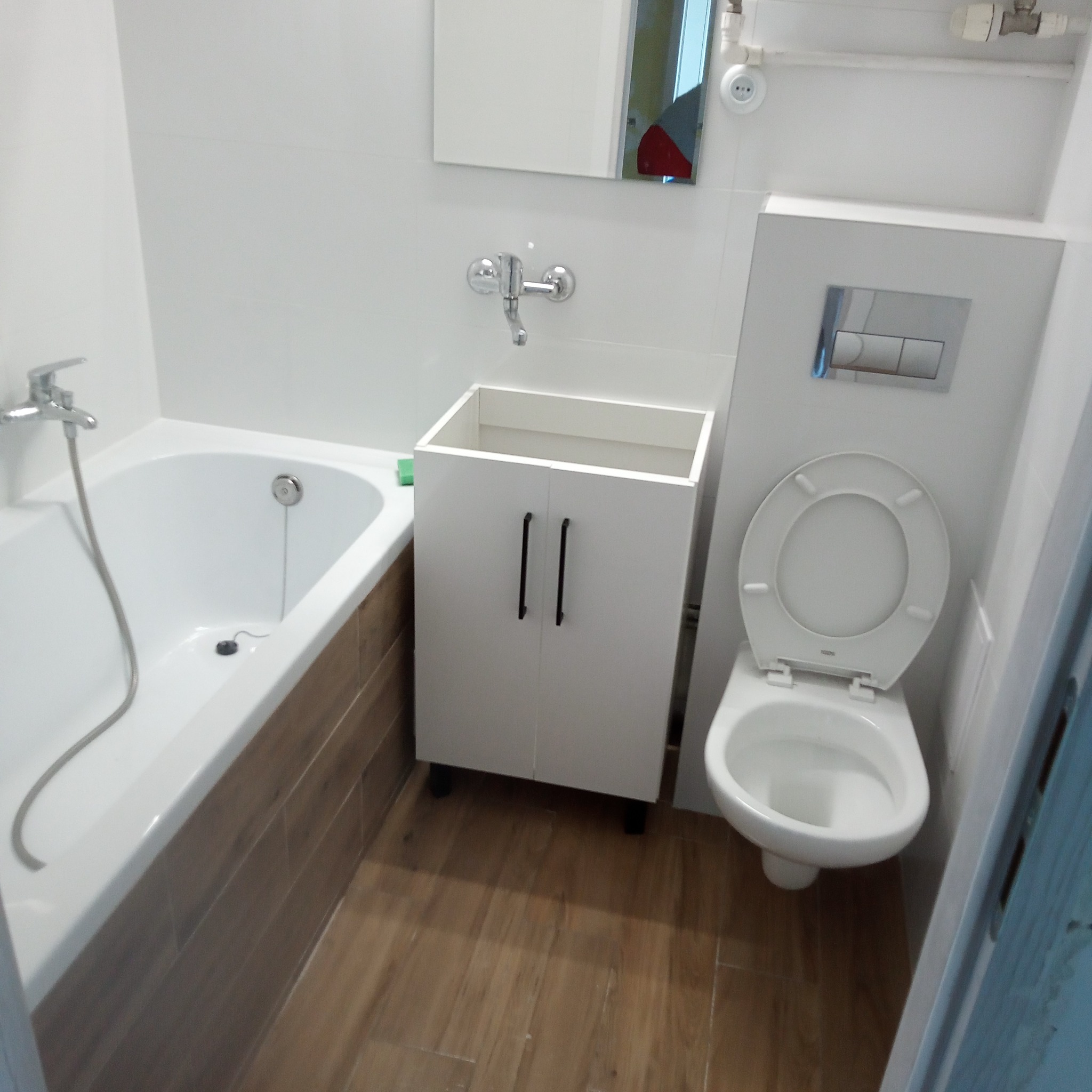 Biała łazienka z wanną obudowaną drewnopodobnymi płytkami, umywalką z szafką i podwieszaną toaletą. Minimalistyczny design, jasne wnętrze.