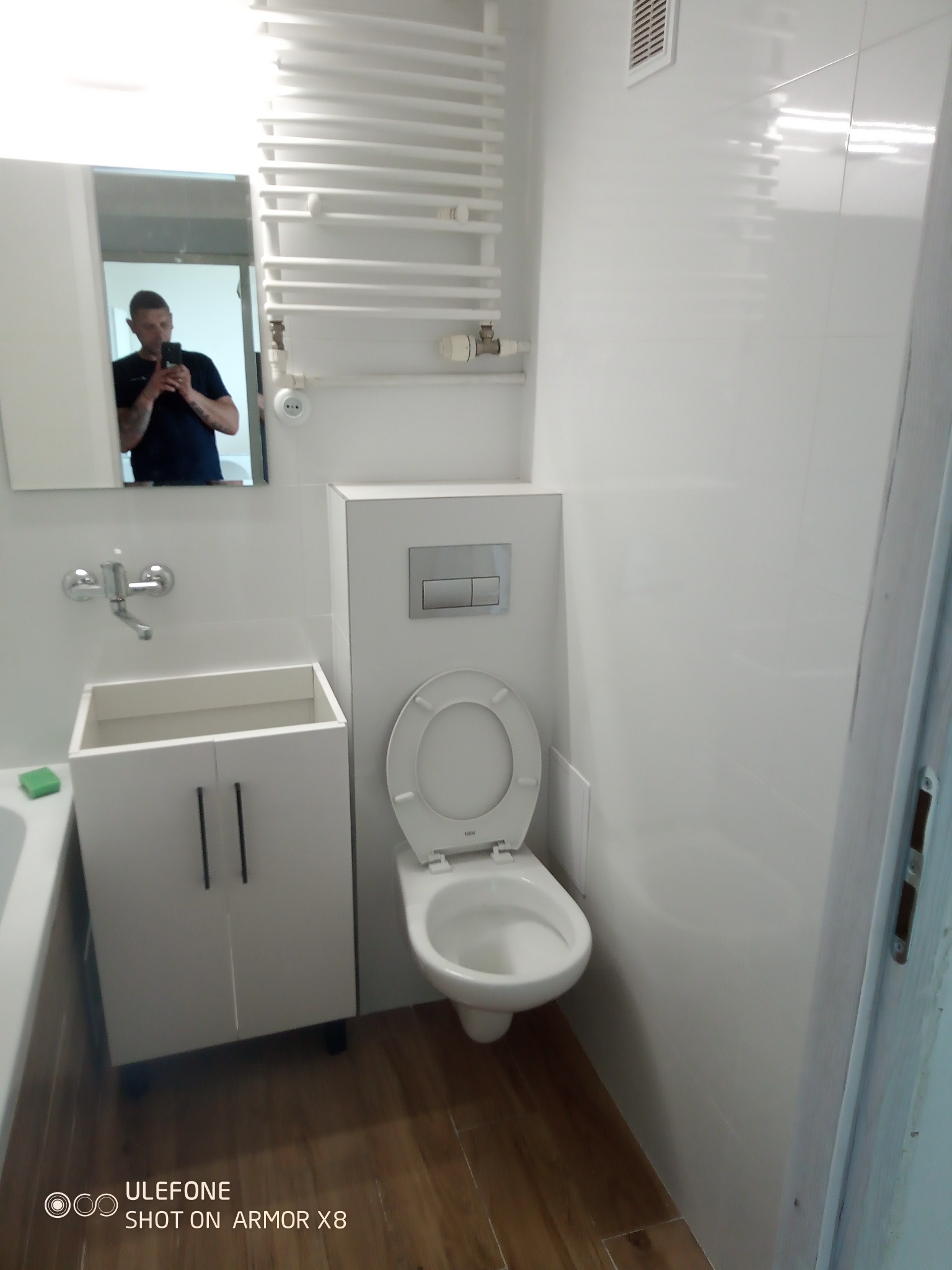 Nowoczesna, biała łazienka po remoncie: podwieszana toaleta, szafka pod umywalką i grzejnik drabinkowy. W lustrze odbija się osoba robiąca zdjęcie.