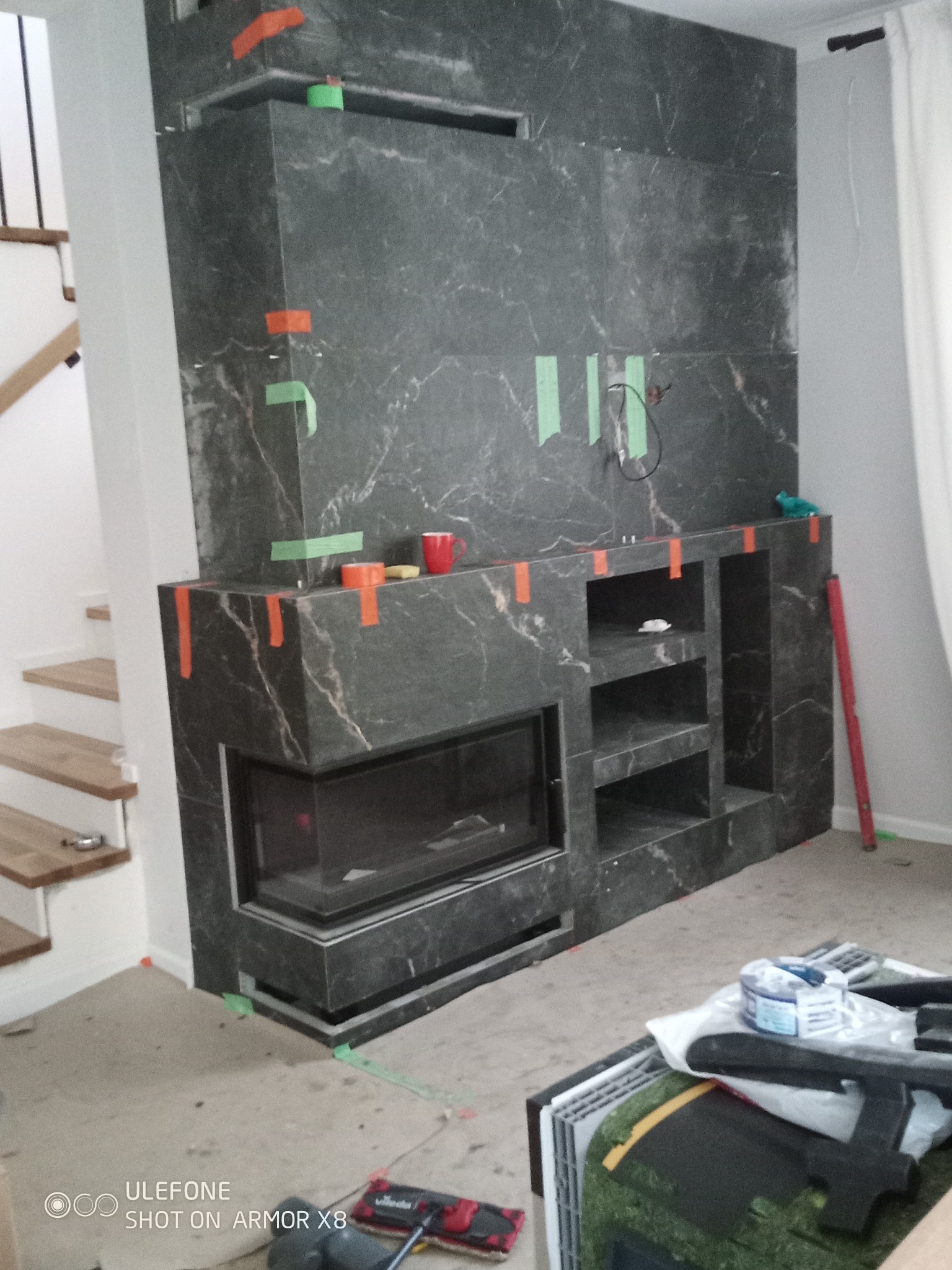 Obudowa kominka z ciemnego marmuru w trakcie montażu, zabezpieczona taśmą malarską. Widoczne narzędzia i poziomica. Schody w tle.