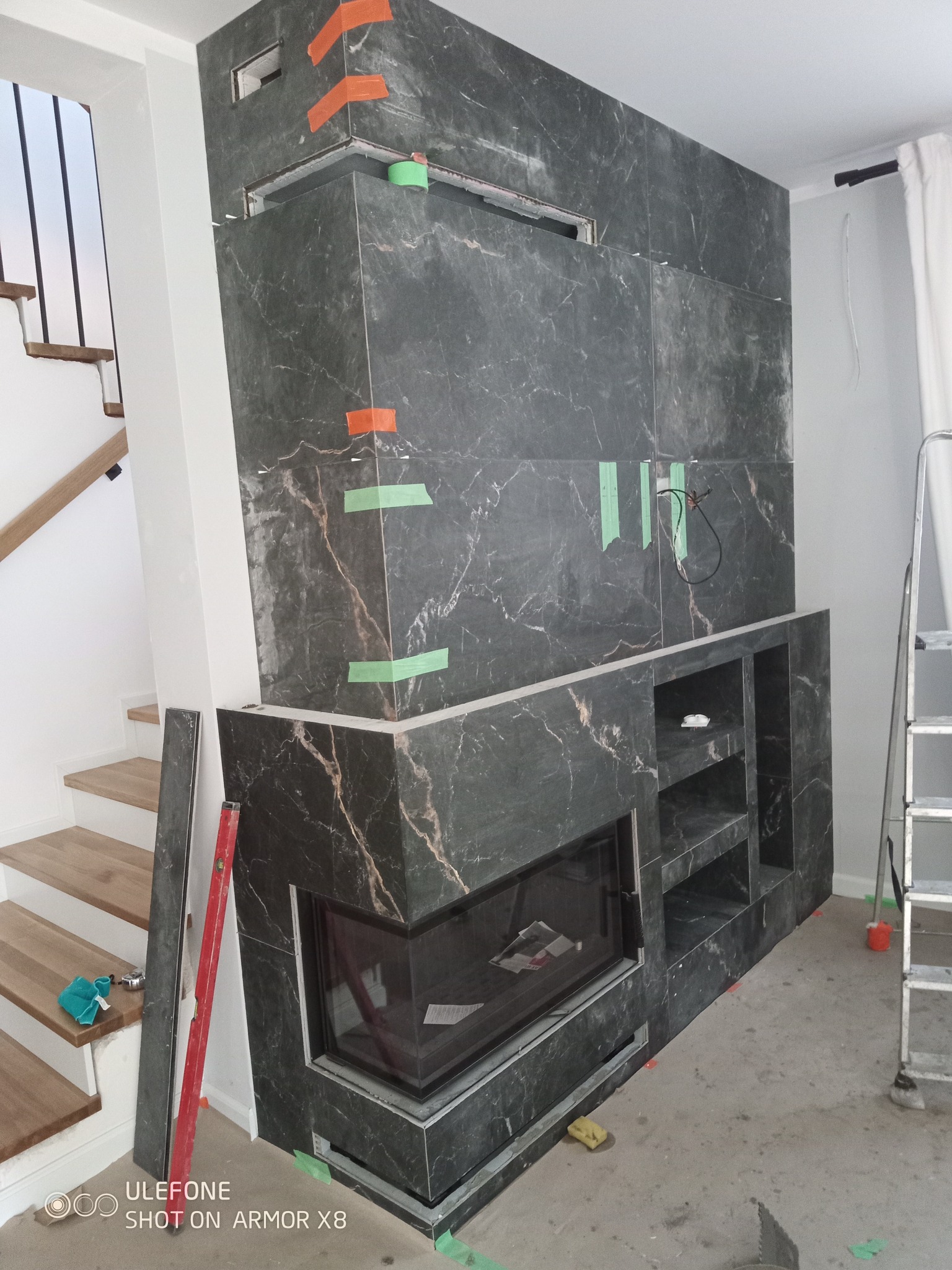 Wnętrze w trakcie wykańczania: obudowa kominka z płyt imitujących marmur, z widocznymi taśmami maskującymi i poziomnicą. Schody w tle dodają perspektywy.