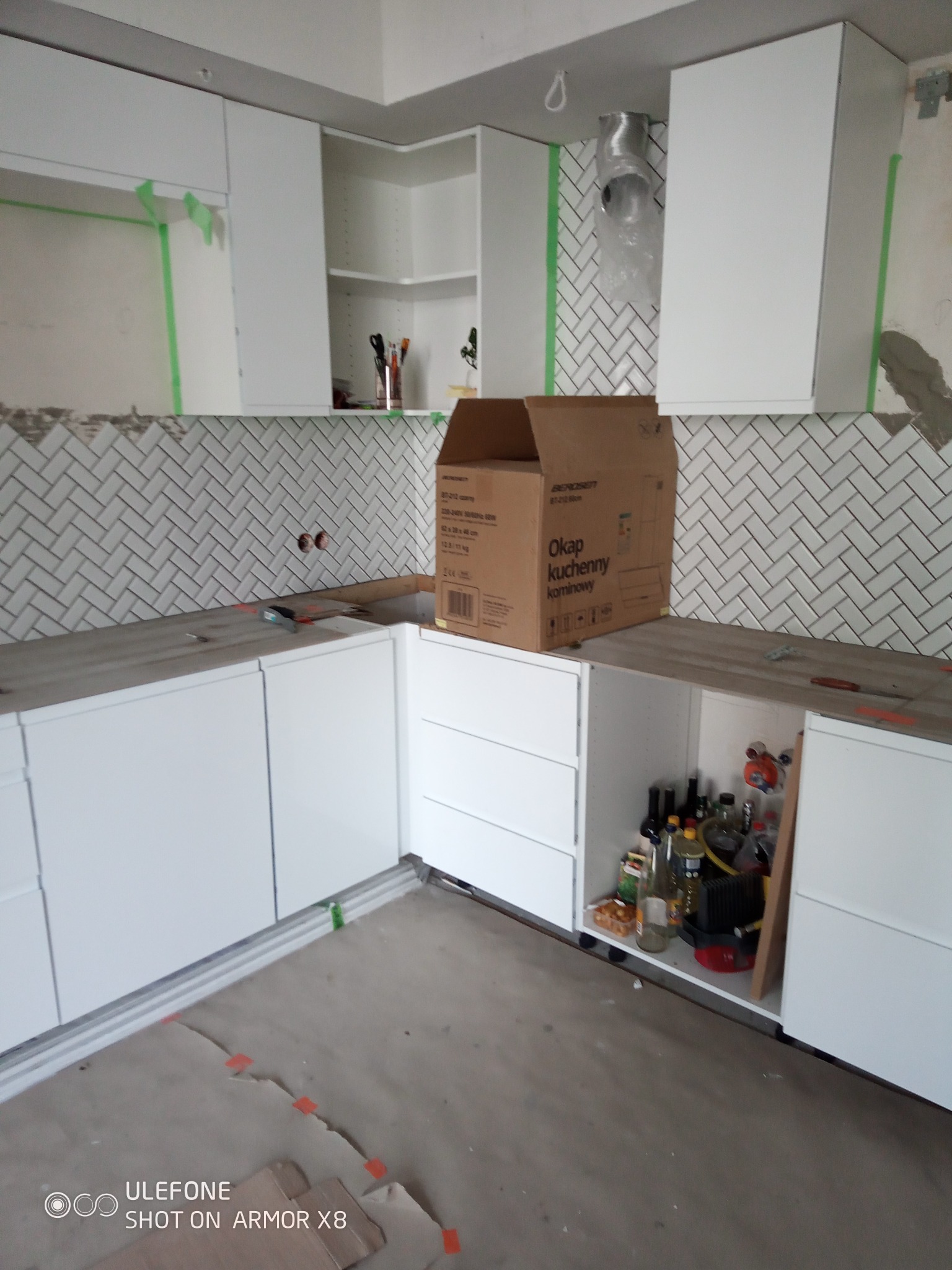 Montaż białych szafek kuchennych i układanie białej glazury w jodełkę. Widoczna kartonowa obudowa okapu i narzędzia na blacie. Etap wykończenia.