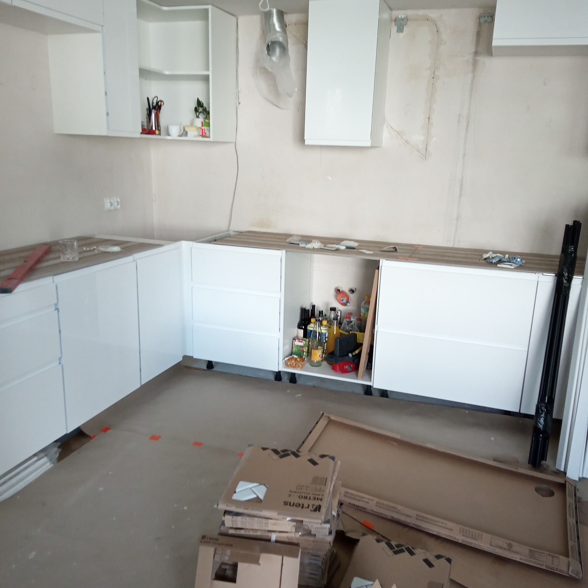 Montaż białych szafek kuchennych z drewnianym blatem na tle surowej ściany. Na podłodze kartony i elementy montażowe. Widoczny fragment instalacji wentylacyjnej.