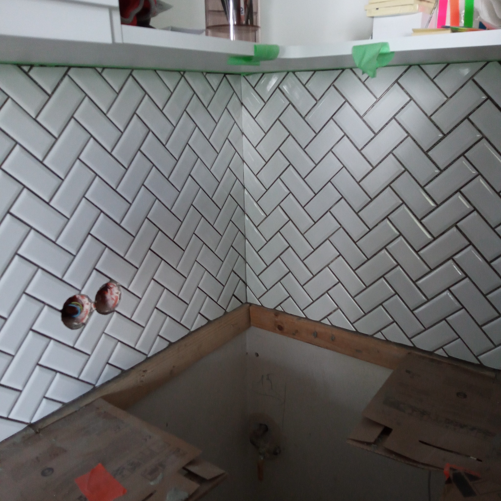 Ukończona ściana z białych, ułożonych w jodełkę płytek, z widocznymi otworami na instalacje i fragmentem niedokończonej zabudowy szafkowej.