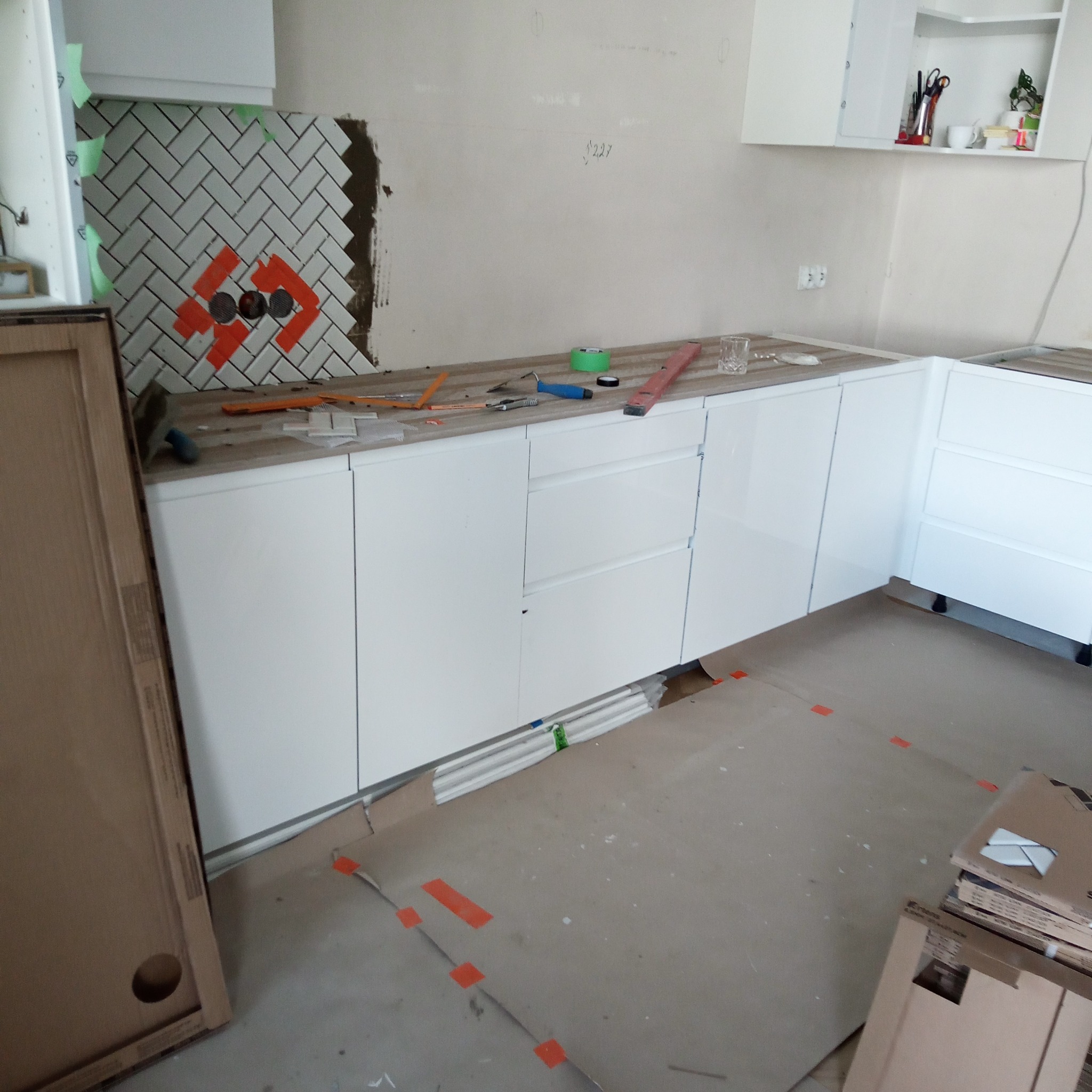 Kuchenny fragment ściany z ułożoną białą glazurą w jodełkę, z widocznymi otworami i pomarańczowymi oznaczeniami. Na blacie narzędzia glazurnicze i nowe, białe szafki.