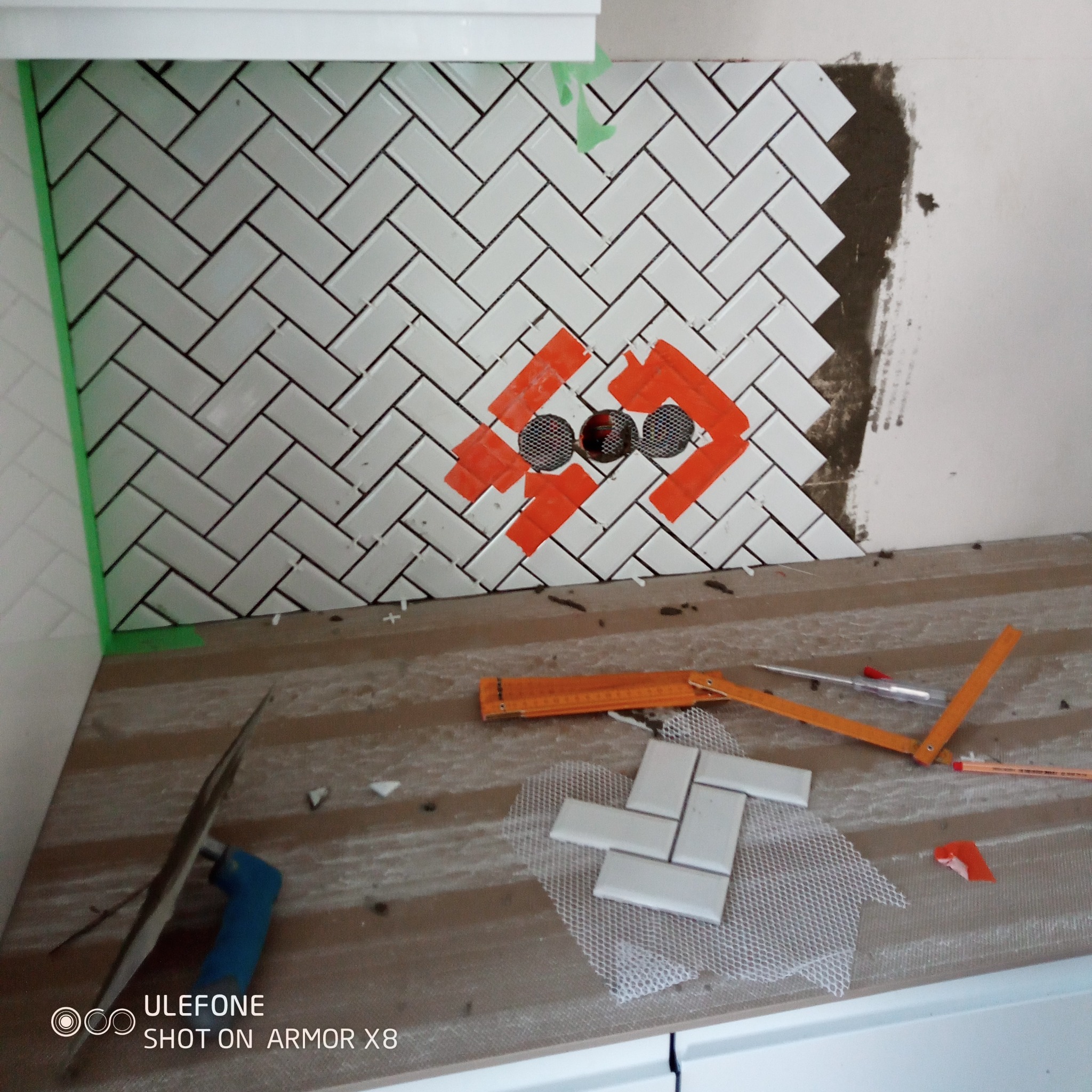 Układanie białej glazury w jodełkę na ścianie z otworem na rury, zabezpieczonym taśmą. Widoczne narzędzia glazurnicze i resztki płytek. Praca w toku.