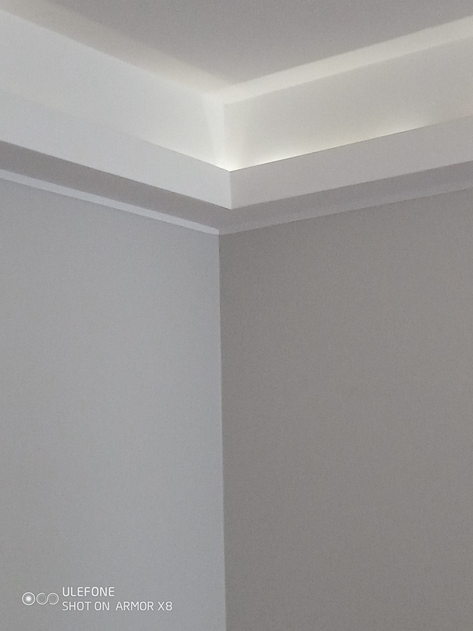 Pomieszczenie po remoncie: Szare ściany i biały sufit z listwami dekoracyjnymi, subtelne oświetlenie LED wzdłuż krawędzi sufitu. Ujęcie z dołu, widoczny róg pokoju.