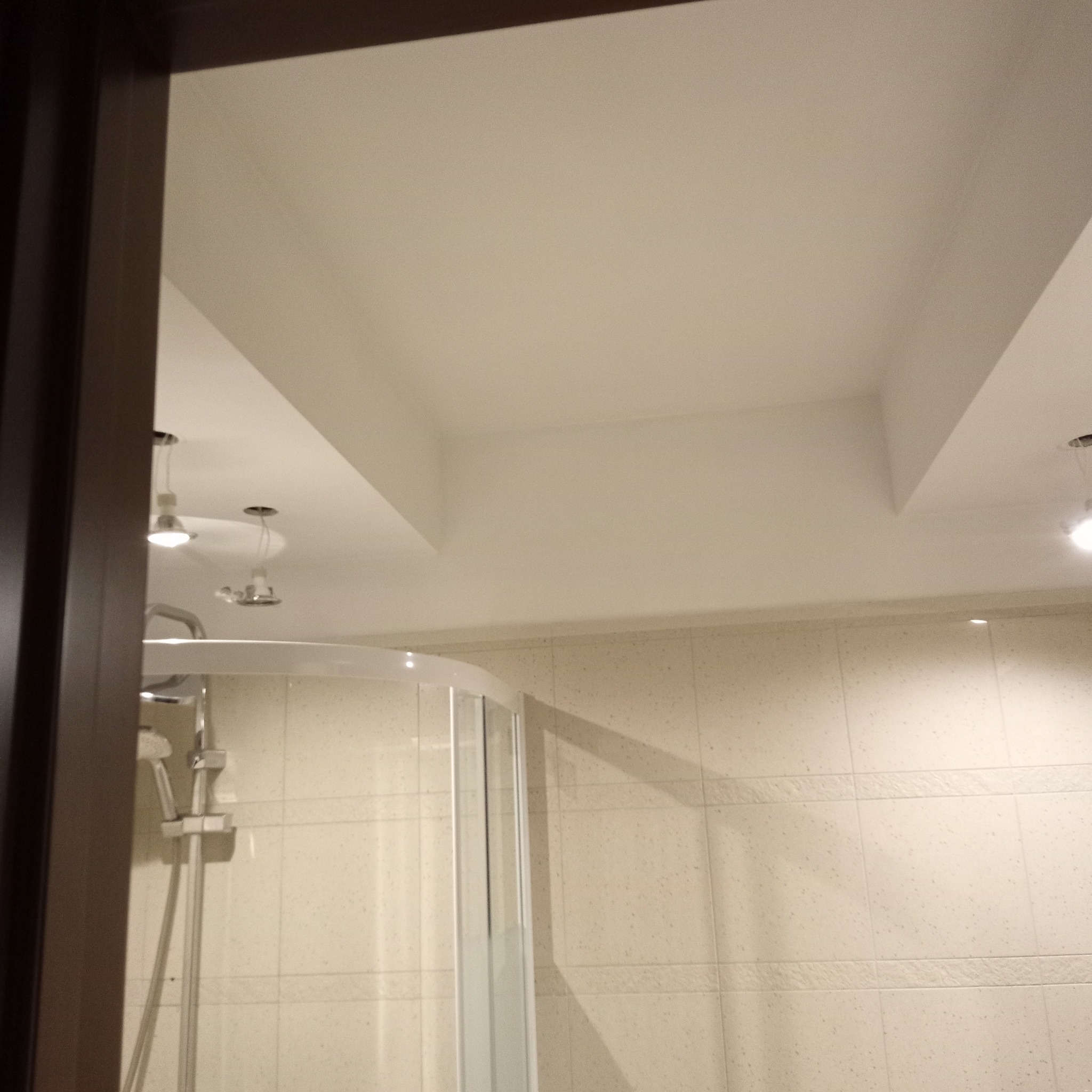 Wykończona łazienka z kabiną prysznicową i beżowymi płytkami. Widoczne oświetlenie sufitowe i fragment ramy drzwi. Ujęcie z boku, z widocznym fragmentem kabiny.