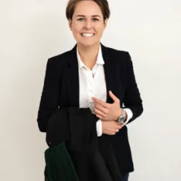 Uśmiechnięta kobieta w białej koszuli i marynarce, trzymająca togę adwokacką z zielonymi wstawkami na tle jasnej ściany. Widoczny zegarek na nadgarstku.