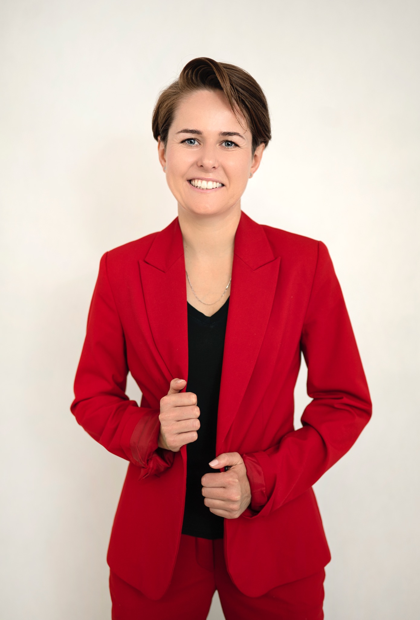 Uśmiechnięta kobieta w czerwonym garniturze, czarnej bluzce i srebrnym naszyjniku, pozująca na jasnym tle. Krótkie, brązowe włosy. Ujęcie portretowe.