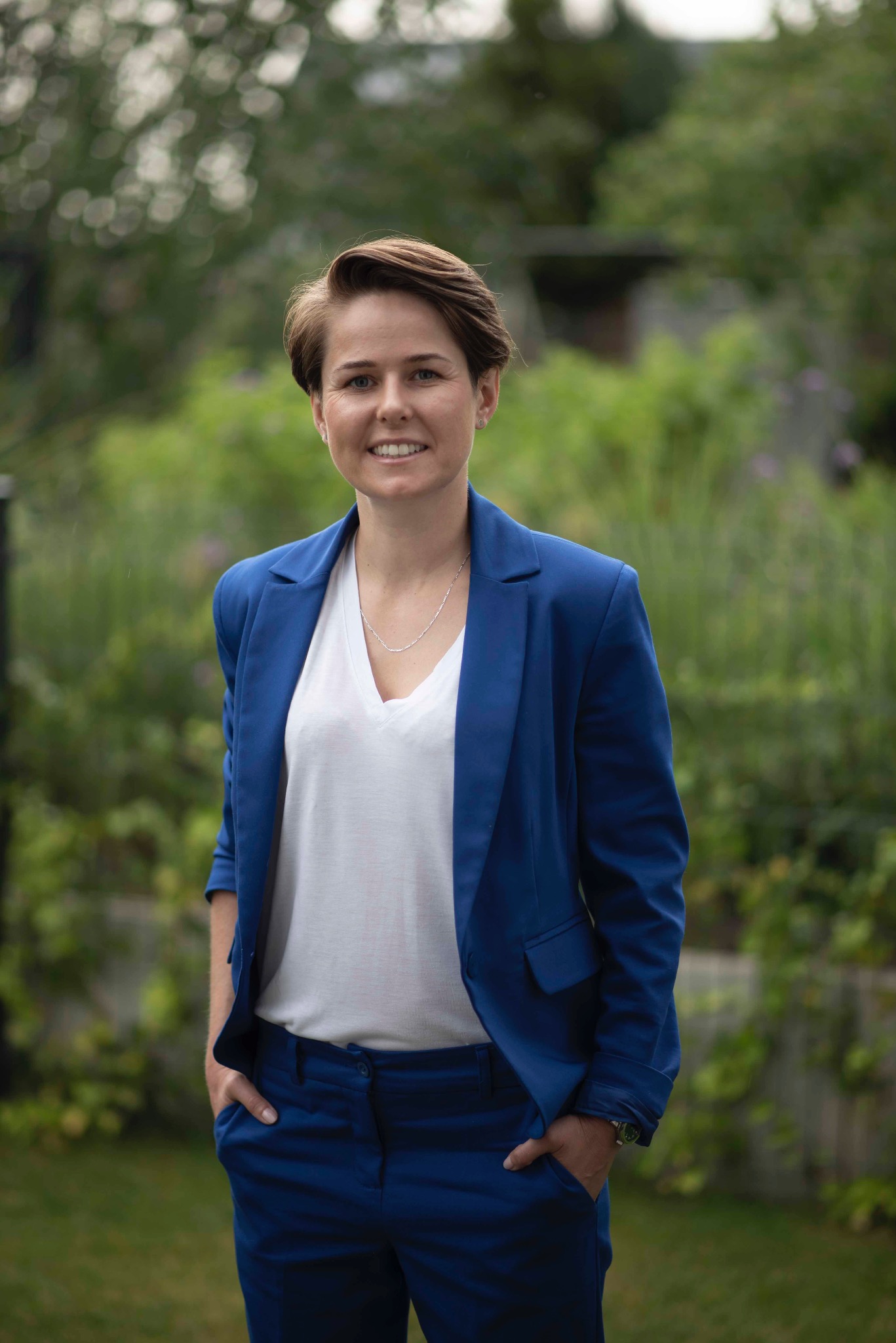 Portret kobiety w niebieskim garniturze, stojącej na tle zieleni. Uśmiech, krótka fryzura. Drobny srebrny łańcuszek na szyi. Ręce w kieszeniach spodni.