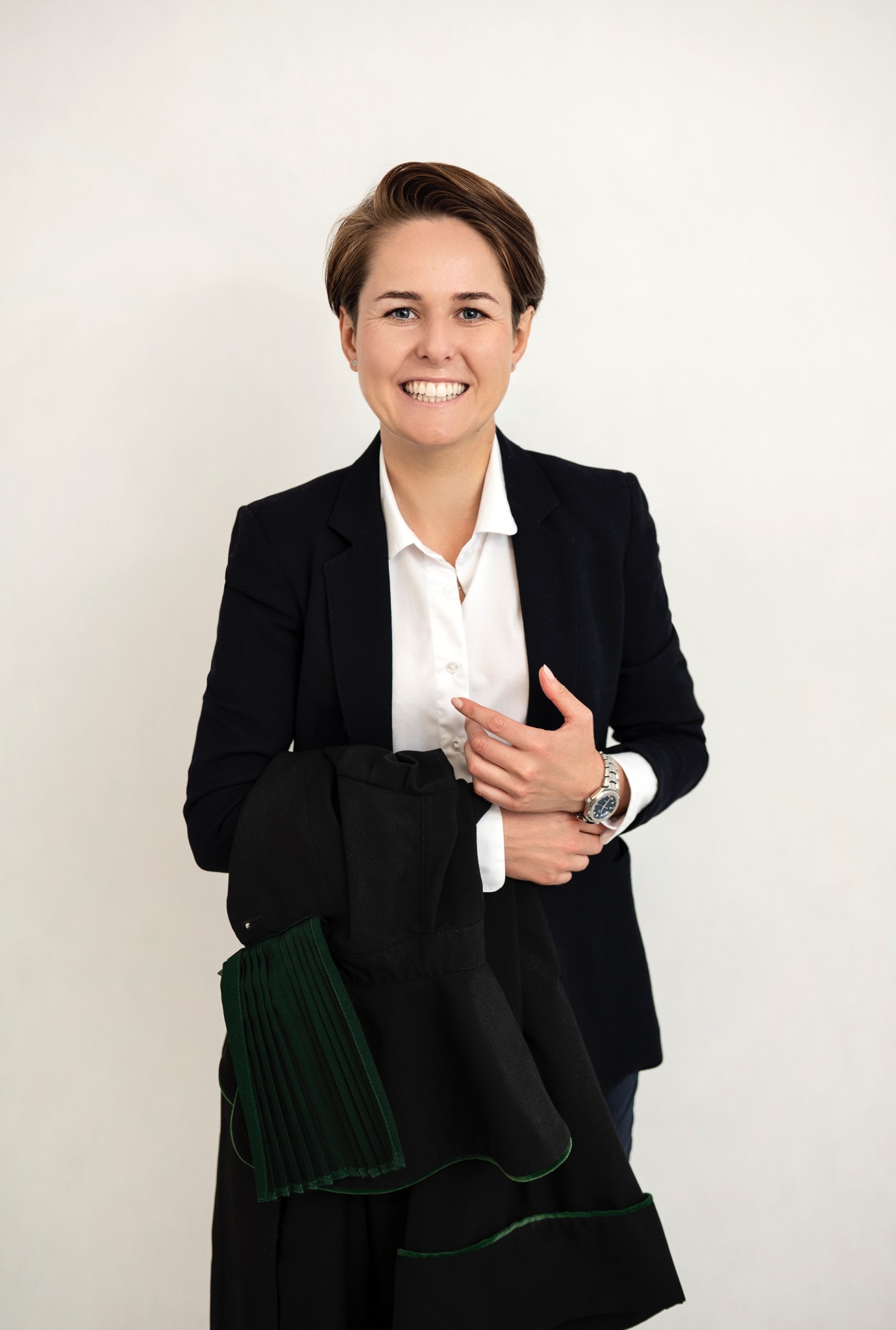 Uśmiechnięta kobieta w białej koszuli i marynarce, trzymająca togę adwokacką z zielonymi wstawkami na tle jasnej ściany. Widoczny zegarek na nadgarstku.