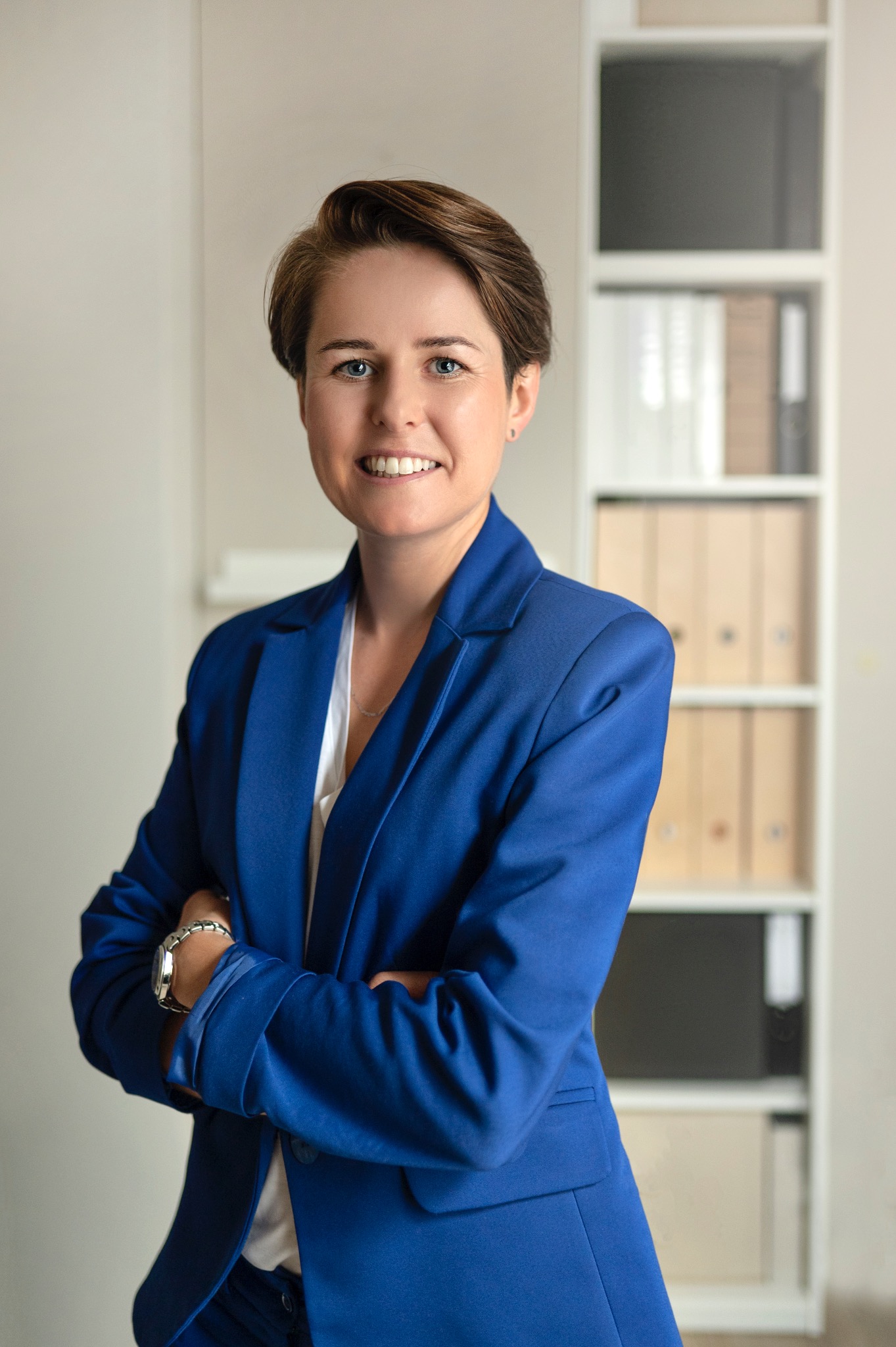 Portret uśmiechniętej kobiety w niebieskim garniturze, z założonymi rękami, na tle regału z segregatorami w jasnym biurze. Profesjonalny wizerunek.