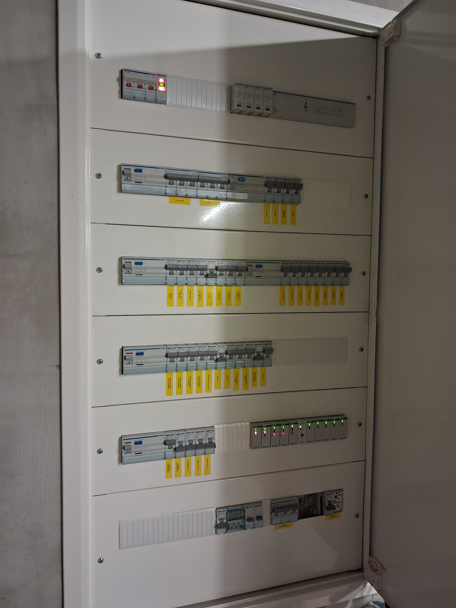 Rozdzielnica elektryczna w obudowie, widoczne zabezpieczenia z żółtymi etykietami i diody sygnalizacyjne. Element instalacji elektrycznej wewnątrz budynku.