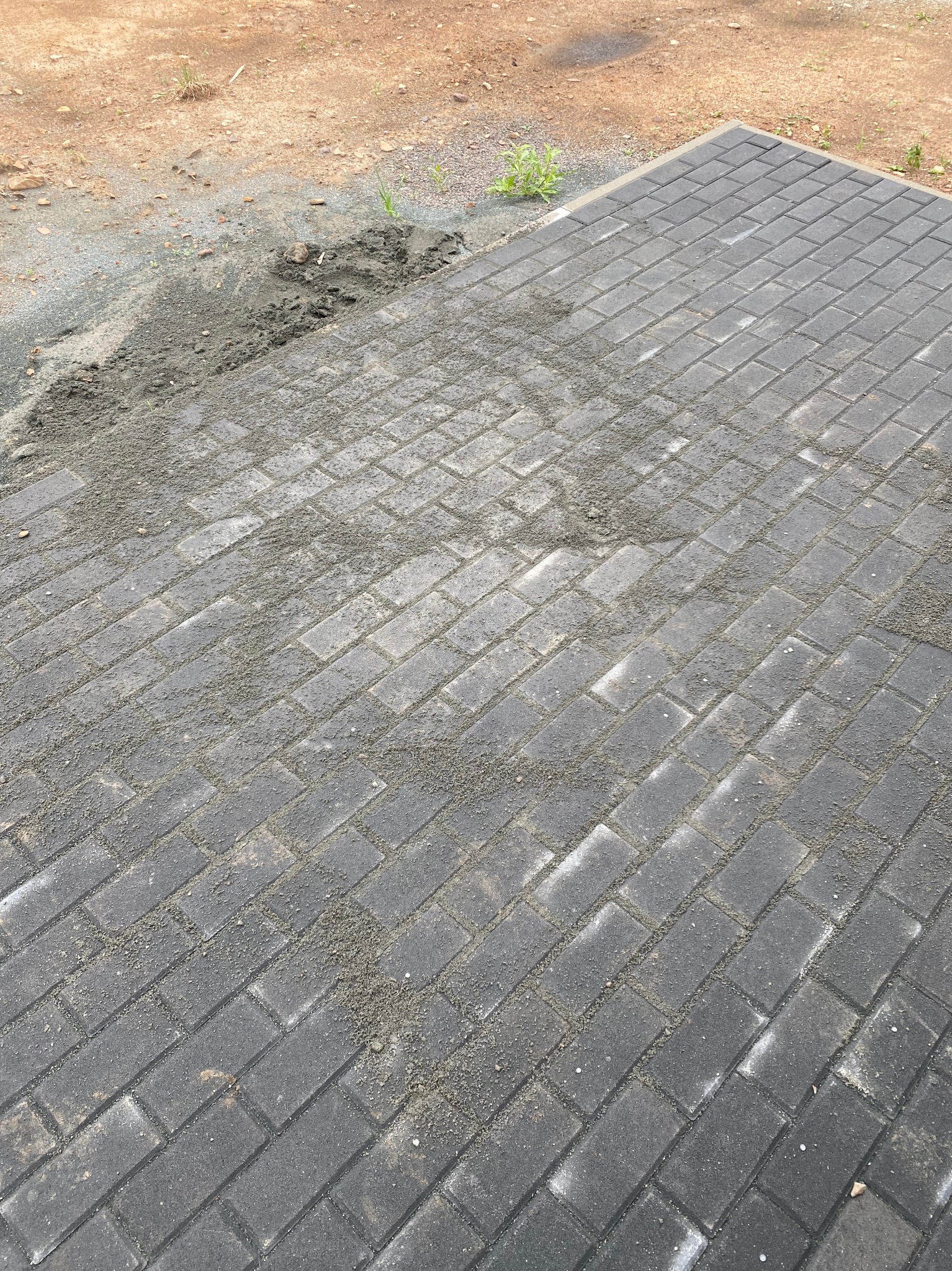 Szara kostka brukowa ułożona w cegiełkę, częściowo zasypana piaskiem, widoczne niedociągnięcia. Tło: ziemia i żwir.