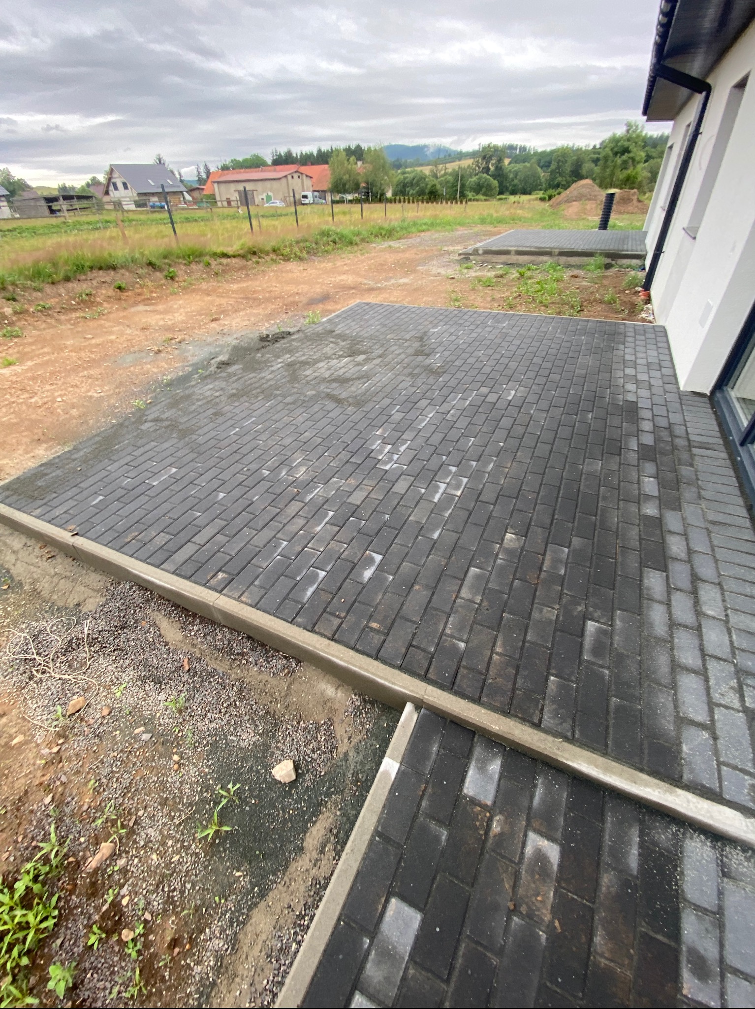 Ułożona, grafitowa kostka brukowa przy domu jednorodzinnym, obrzeża betonowe. Widok na wiejską okolicę w tle.
