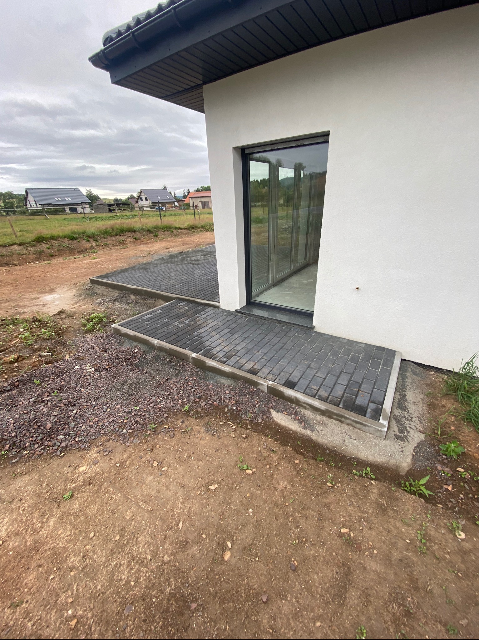 Ułożona kostka brukowa przed wejściem do nowoczesnego domu z dużym oknem. Widoczne niedokończone prace ziemne i fragmenty trawnika w tle.