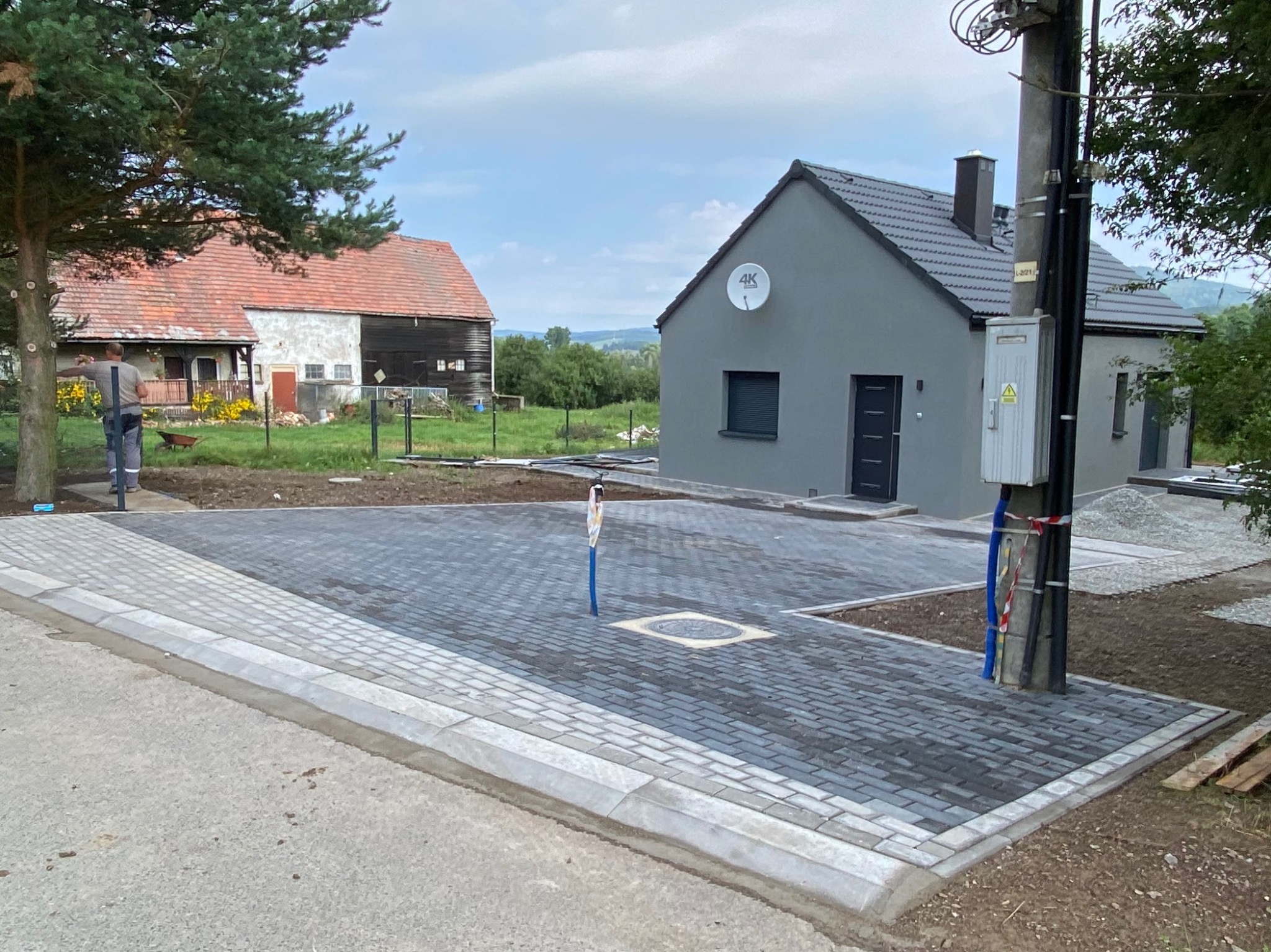 Ułożona kostka brukowa w odcieniach szarości, obrzeża betonowe, widok na dom i słup energetyczny. Częściowo zagospodarowany teren.