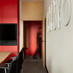 Nowoczesne biuro z czerwonymi szafkami, drewnianymi elementami i dekoracją ścian w postaci białych rakiet tenisowych. Osoba przechodzi przez otwarte drzwi.