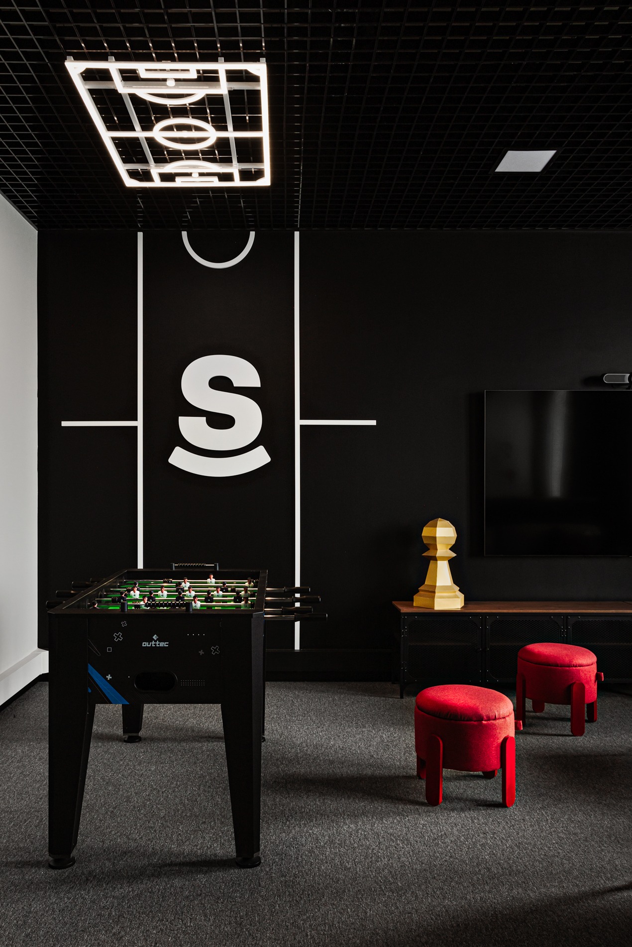 Stylowe biuro z czarną ścianą, białym logo 'S' i motywem boiska. Stół do piłkarzyków, czerwone pufy, szara wykładzina. Nietypowa lampa sufitowa w kształcie boiska.