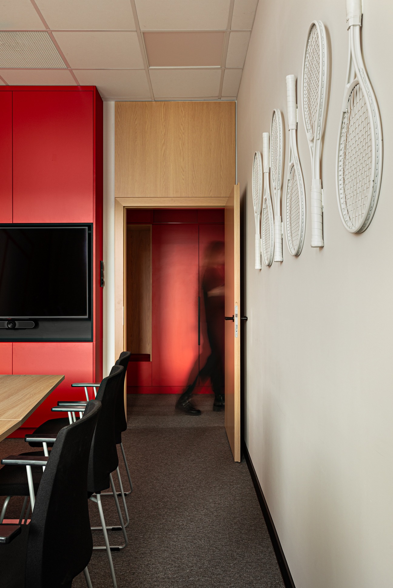 Nowoczesne biuro z czerwonymi szafkami, drewnianymi elementami i dekoracją ścian w postaci białych rakiet tenisowych. Osoba przechodzi przez otwarte drzwi.