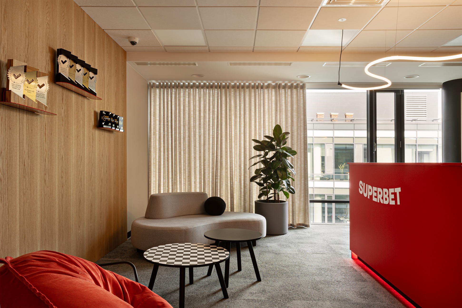 Nowoczesna aranżacja biura z recepcją Superbet, drewnianą ścianą i nagrodami. Wygodna sofa i stoliki kawowe tworzą strefę relaksu z rośliną doniczkową.