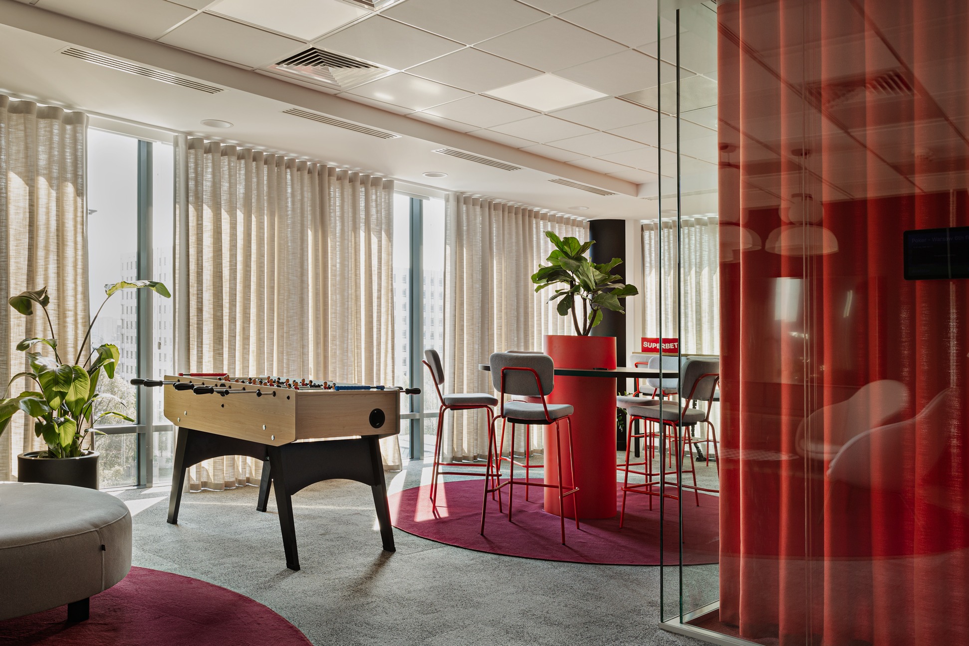 Nowoczesna aranżacja biura z piłkarzykami, wysokimi krzesłami i roślinami doniczkowymi. Widoczne duże okna z jasnymi zasłonami, tworzące przestrzeń relaksu.