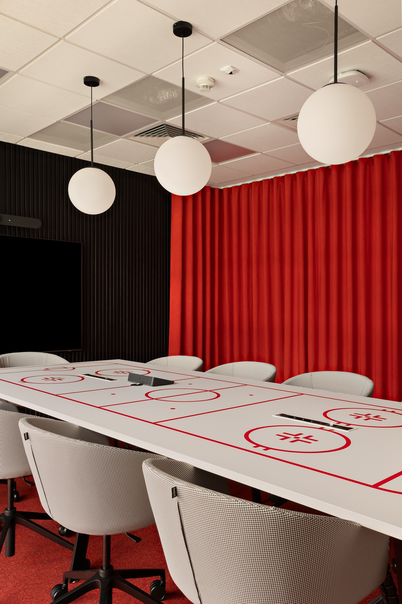 Sala konferencyjna z długim stołem w hokejowym stylu, otoczonym krzesłami w pepitkę. Czerwone zasłony i czarna ściana z TV tworzą kontrast.