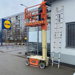 Parsbud Sp.z.o.oo - Pracownik na podnośniku koszowym dokonuje naprawy/montażu na elewacji budynku z logo Lidl. Obok drabina. Ujęcie z dołu, na tle bloku mieszkalnego.