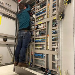 Parsbud Sp.z.o.oo - Elektryk na podeście, w trakcie pracy przy rozbudowanej szafie sterowniczej z licznymi przewodami i elementami elektrycznymi. Widok z dołu, wnętrze pomieszczenia.