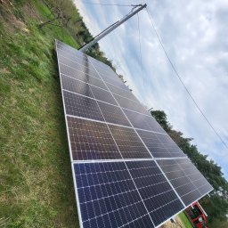 Parsbud Sp.z.o.oo - Panel fotowoltaiczny na trawie, widok z dołu. Słup energetyczny w tle. Instalacja paneli słonecznych w otoczeniu zieleni i błękitnego nieba z chmurami.