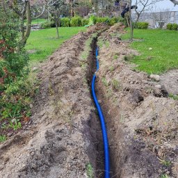 Parsbud Sp.z.o.oo - Niebieska rura karbowana w wykopie na trawniku, prawdopodobnie instalacja podziemna. Widok z góry na tle domu i drzew.