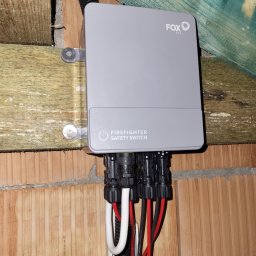 Parsbud Sp.z.o.oo - Szary wyłącznik bezpieczeństwa pożarowego marki Fox ESS, zamontowany na drewnianej konstrukcji i ceglanej ścianie, z widocznymi kablami i złączkami.
