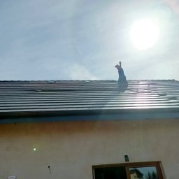 Parsbud Sp.z.o.oo - Montaż paneli fotowoltaicznych na dachu domu z drabiny. Pracownik w kombinezonie na tle jasnego nieba i słońca. Widok z dołu na dachówkę.