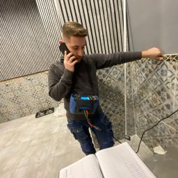 Parsbud Sp.z.o.oo - Elektryk w trakcie pracy: rozmawia przez telefon, opierając się o ścianę z wzorzystymi płytkami, z miernikiem na pasie i notatnikiem na blacie.
