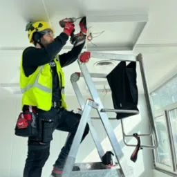 Elektryk na drabinie montuje oświetlenie sufitowe w jasnym pomieszczeniu. Widoczne narzędzia i przewody. Pracownik w kasku i kamizelce odblaskowej.