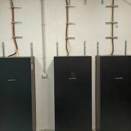 Parsbud Sp.z.o.oo - Trzy czarne akumulatory energii Sonnen podłączone do instalacji elektrycznej na ścianie z widocznymi kablami i uchwytami montażowymi.