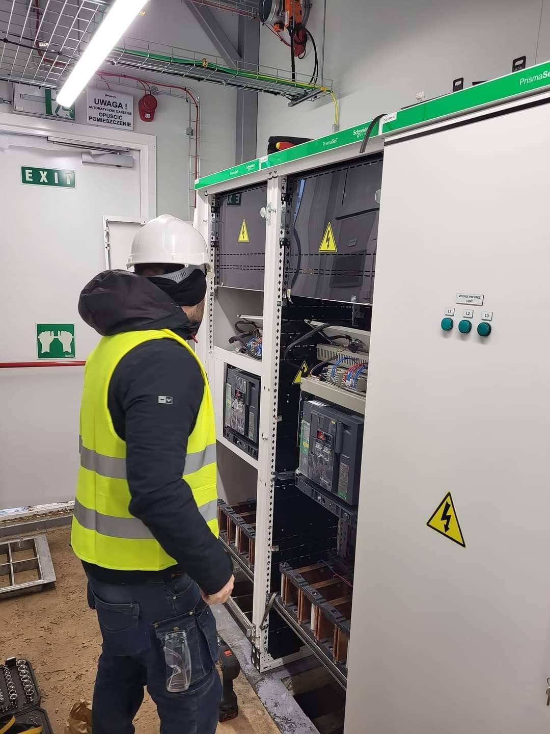 Elektryk w kasku i kamizelce BHP przy rozdzielnicy elektrycznej PrismaSeT. Widoczne kable, bezpieczniki i oznaczenia ostrzegawcze. Elementy instalacji w tle.