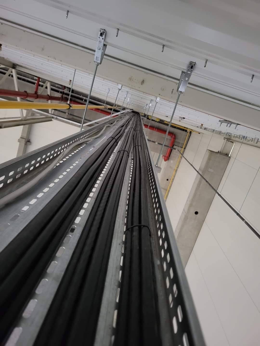 Rzut z dołu na koryto kablowe z czarnymi przewodami, mocowane do sufitu za pomocą metalowych uchwytów. Widoczne rury instalacyjne w tle.